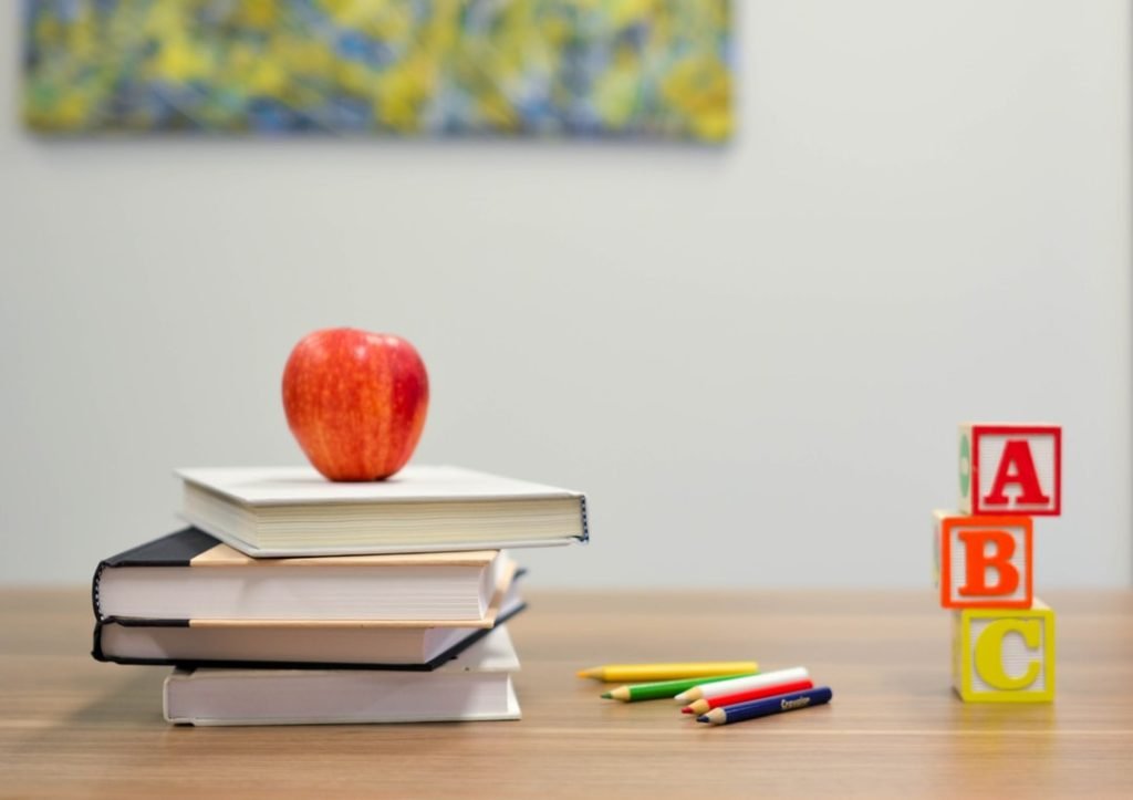 A SPED teacher's desk has an apple sitting on four stacked books, a few colored pencils, and some ABC blocks on it.