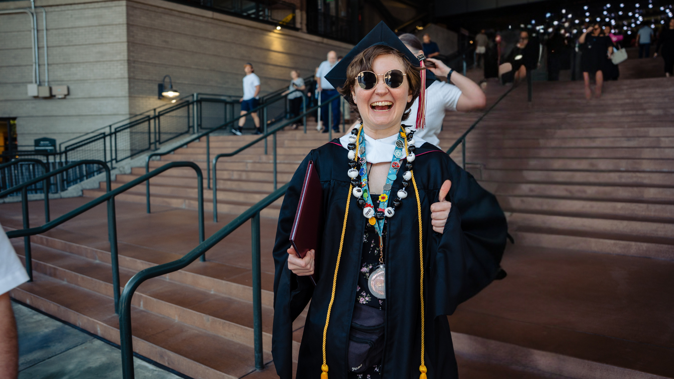 graduating student in regalia with sunglasses on outside of commencement smiling and giving thumbs up to tuition reimbursement