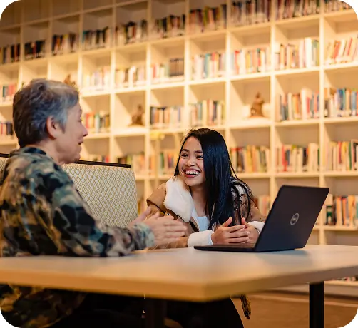 Two students having a conversation at a table in a library.